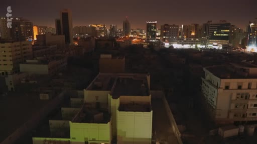 Vue aérienne nocturne d'un paysage urbain dense avec des gratte-ciels illuminés et des zones résidentielles au premier plan, illustrant l'urbanisation nocturne.