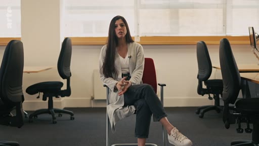 Une femme aux longs cheveux bruns est assise sur une chaise dans un bureau moderne et lumineux, répondant à une interview. Elle porte un gilet clair et un jean, et est entourée de chaises de bureau vides.