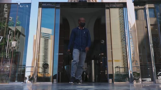 Un homme portant un masque sort d'un immeuble de bureaux moderne à Dubaï, aux Émirats arabes unis. L'architecture contemporaine et les reflets sur les façades vitrées caractérisent le paysage urbain de la ville.