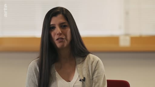 Une femme aux longs cheveux bruns, vêtue d'un gilet gris sur un haut blanc, est filmée en plan moyen lors d'une interview télévisée. Elle s'adresse directement à la caméra dans un cadre intérieur sobre.