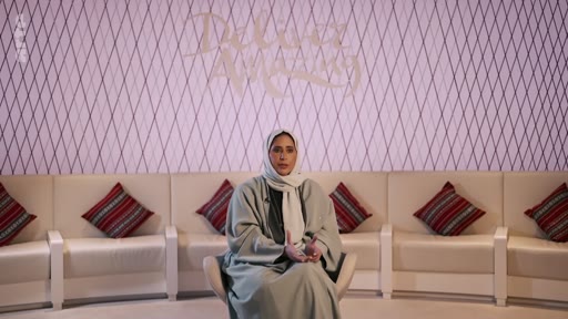 Une femme portant un hijab et une abaya est assise sur un canapé blanc devant un mur décoré avec l'inscription 'Deliver Amazing' lors d'une interview télévisée.