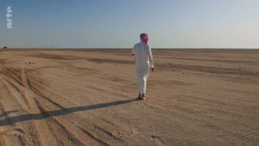 Un homme portant une thobe blanche et un keffieh marche seul dans une étendue désertique aride sous un ciel clair. Cette image illustre la solitude et l'immensité des paysages désertiques du Moyen-Orient.
