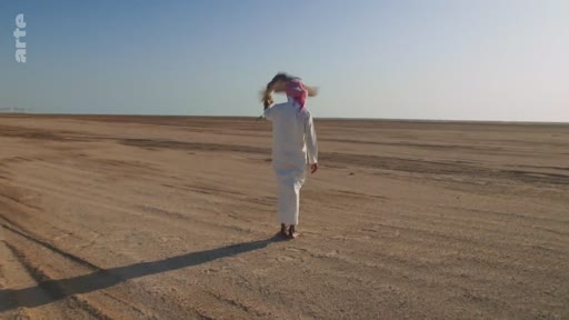 Un homme portant une thobe blanche et un keffieh marche pieds nus dans une étendue désertique aride sous un ciel clair. Cette image illustre la solitude et la vie dans les régions désertiques du Moyen-Orient.