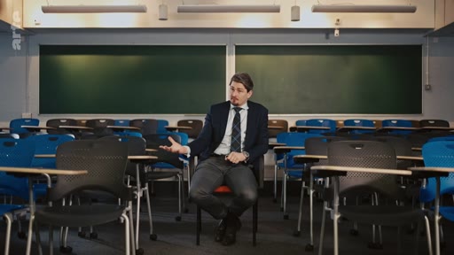 Un homme en costume, assis sur une chaise au centre d'une salle de classe vide avec des tableaux noirs en arrière-plan, s'exprime lors d'une interview.
