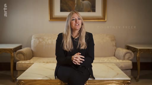 La chercheuse Courtney Freer est filmée en plan moyen lors d'une interview, assise devant un canapé dans un salon formel. Elle s'exprime sur des sujets d'actualité ou de recherche dans le cadre d'un reportage télévisé.