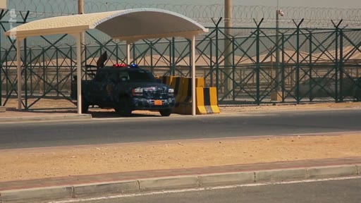Un véhicule de police avec des gyrophares est stationné sous un abri devant une clôture grillagée surmontée de barbelés, dans une zone désertique. Cette image illustre des mesures de sécurité renforcées ou une zone de haute surveillance.