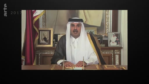 L'émir du Qatar, Tamim ben Hamad Al Thani, est filmé en plan moyen derrière un bureau lors d'une allocution officielle en 2017. Il porte la tenue traditionnelle qatarienne devant le drapeau national et des portraits officiels.