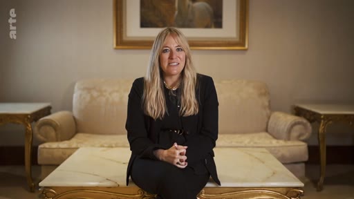 Une femme blonde, vêtue d'une veste noire, est assise sur une table basse dans un salon luxueux, face à la caméra pour une interview.
