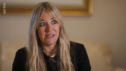 Une femme blonde aux cheveux longs s'exprime face caméra lors d'une interview télévisée, portant un haut noir et un collier doré.