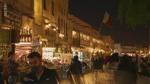 Vue nocturne animée du Souq Waqif à Doha, au Qatar, avec des passants et des boutiques traditionnelles. Le drapeau du Qatar est visible au-dessus des bâtiments historiques illuminés.