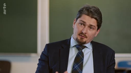 Un homme en costume sombre et cravate à motifs s'exprime face caméra lors d'une interview, avec un tableau noir en arrière-plan.