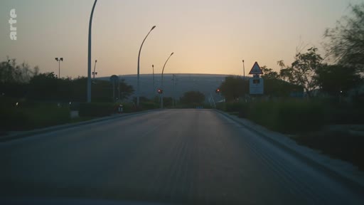 Vue d'une route asphaltée menant vers un grand stade au loin, sous une lumière de fin de journée ou de crépuscule. Le paysage est dégagé avec des lampadaires le long de la voie.