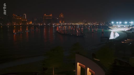 Vue panoramique nocturne de la baie de Doha au Qatar, mettant en évidence les gratte-ciels illuminés et les bateaux traditionnels amarrés sur l'eau. Cette image illustre le développement urbain et le paysage côtier de la capitale qatarie.