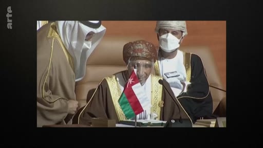Le sultan Haitham ben Tariq d'Oman, portant un masque et une visière de protection, est photographié lors d'un événement officiel avec le drapeau omanais devant lui. Il est entouré de dignitaires portant des tenues traditionnelles dans un contexte de protocole diplomatique.