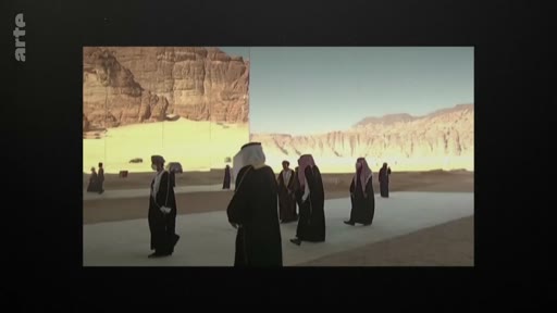 Un groupe d'hommes vêtus de tenues traditionnelles, incluant des thobes et des ghutras, marche sur une surface plane devant des formations rocheuses dans un paysage désertique, évoquant une scène en Arabie saoudite.