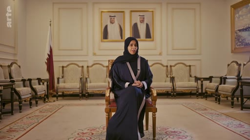 Une femme portant une abaya et un hijab est assise dans un salon officiel au Qatar, avec le drapeau du Qatar et des portraits de dirigeants en arrière-plan. Cette scène illustre une interview formelle dans un contexte diplomatique ou institutionnel qatari.