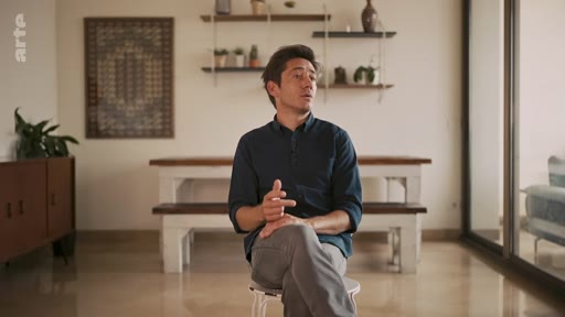 Un homme est assis sur un tabouret dans un salon lumineux, en train de parler lors d'une interview. Il porte une chemise bleu foncé et un pantalon gris, avec un décor minimaliste en arrière-plan.