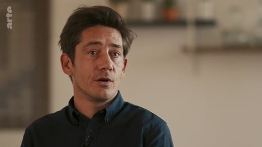 Un homme s'exprime face caméra lors d'une interview dans un cadre intérieur. Il porte une chemise sombre et semble participer à un documentaire ou une émission de télévision.