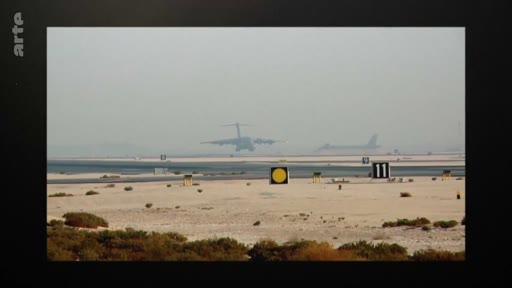 Un avion de transport militaire, probablement un C-17 Globemaster, effectue un atterrissage sur une piste d'aéroport située dans un environnement désertique. La scène montre l'activité aérienne sur une base militaire ou un aéroport en zone aride.