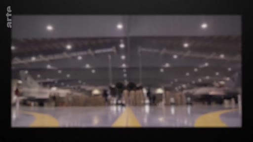 Vue d'ensemble d'un hangar militaire abritant plusieurs avions de chasse, avec des techniciens travaillant à proximité des appareils. L'image illustre les opérations de maintenance ou de préparation au sein d'une base aérienne.