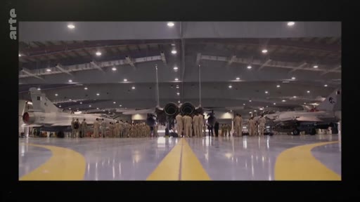 Un groupe de militaires en uniforme est rassemblé dans un grand hangar aéronautique devant plusieurs avions de chasse, dont un F-15 Eagle au centre. Cette scène illustre les opérations de maintenance ou de préparation au sein d'une base aérienne militaire.