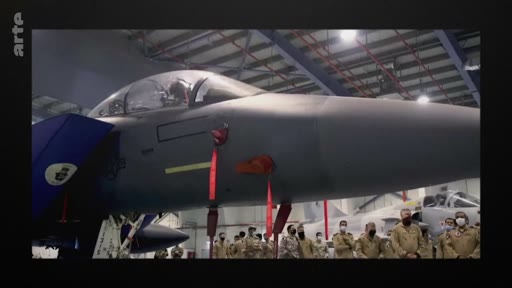 Des militaires en uniforme, certains portant des masques, se tiennent devant un avion de chasse stationné dans un hangar technique. La scène montre une inspection ou une présentation de matériel aéronautique militaire.