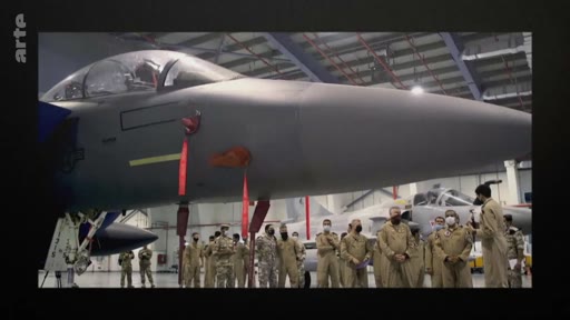 Un groupe de militaires en tenue de vol examine un avion de chasse, probablement un F-15, à l'intérieur d'un hangar d'entretien. La scène montre une inspection technique ou une présentation de matériel aéronautique militaire.