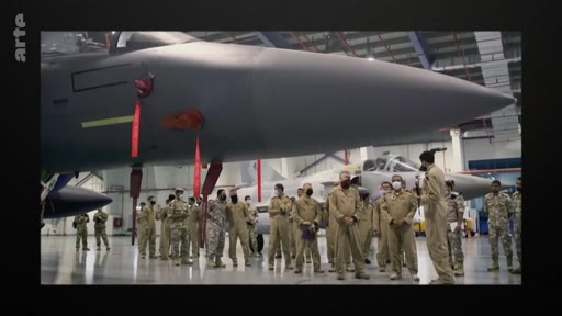 Un groupe de militaires en tenue de vol beige se tient devant un avion de chasse dans un hangar, probablement lors d'une visite ou d'une séance d'information. La scène montre des pilotes et du personnel technique observant un appareil de combat.