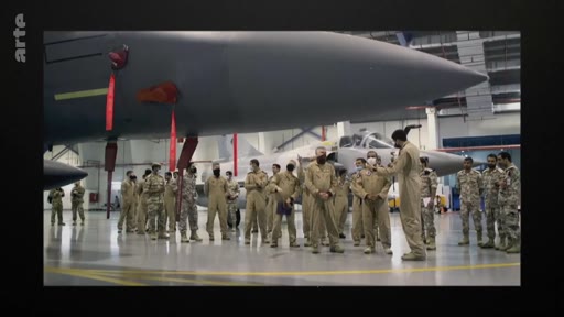 Des pilotes et du personnel militaire en tenue de vol beige sont rassemblés dans un hangar aéronautique pour un briefing devant un avion de combat de type Rafale. La scène montre une préparation opérationnelle au sein d'une base aérienne.