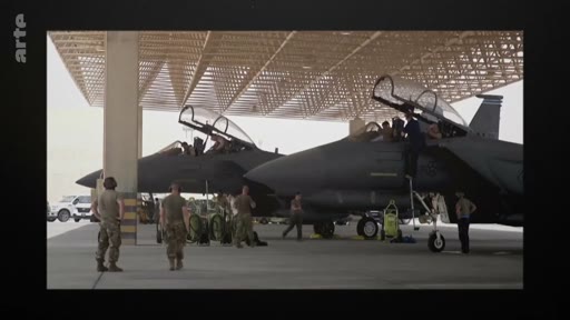 Des militaires en uniforme effectuent la maintenance et la préparation de deux avions de chasse sous un hangar sur une base aérienne. La scène montre l'activité opérationnelle autour des appareils de combat.