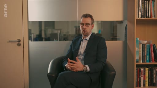 Un homme portant des lunettes et un costume sombre est assis dans un fauteuil, les mains jointes, lors d'une interview télévisée dans un bureau. Il s'exprime face caméra dans un cadre professionnel.