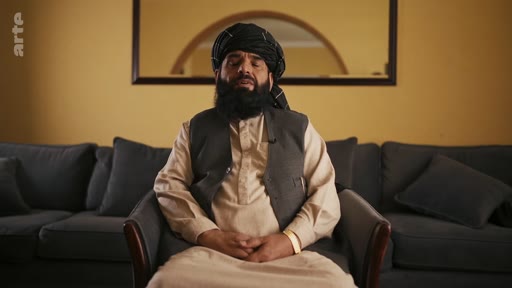 Un homme barbu portant un turban traditionnel est assis dans un fauteuil lors d'une interview. Il est filmé en plan moyen dans un intérieur domestique avec un canapé en arrière-plan.
