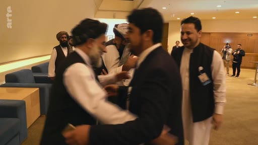 Des membres de la délégation talibane, vêtus de tenues traditionnelles, échangent des salutations avec des officiels lors d'une réunion diplomatique internationale. La scène montre des interactions formelles dans un cadre de conférence.