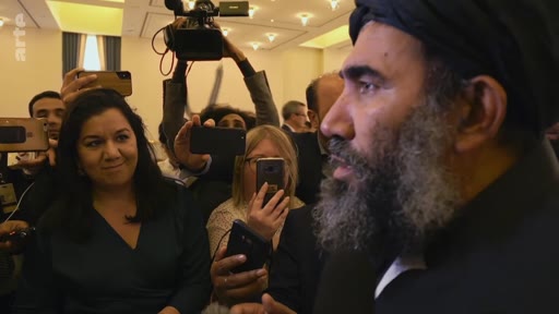 Un homme barbu portant un turban noir est filmé et photographié par un groupe de journalistes lors d'une conférence de presse ou d'un événement public. La scène montre une forte présence médiatique avec plusieurs personnes tenant des smartphones et des caméras pour capturer l'intervention de l'homme.