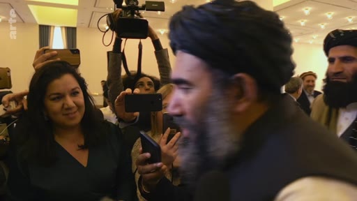 Des journalistes et des photographes entourent des membres des Talibans, dont un homme barbu portant un turban, lors d'une conférence de presse. La scène montre une forte présence médiatique autour des représentants talibans.