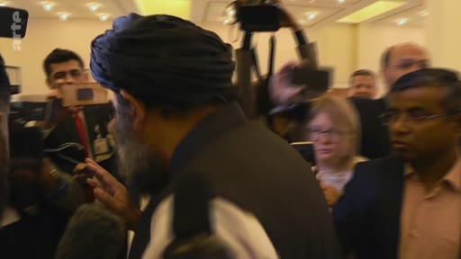Un homme portant un turban sombre est entouré par une foule de journalistes et de cameramen lors d'un événement public. La scène montre une forte présence médiatique autour de cette figure centrale.