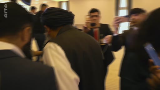 Un homme portant un turban traditionnel est entouré par une foule de journalistes et de caméramans dans un espace intérieur, probablement lors d'une conférence de presse ou d'un événement officiel. La scène montre une forte activité médiatique autour de cette personnalité.