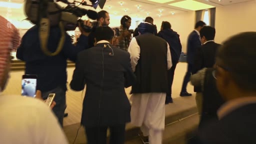 Un groupe de personnes, incluant un homme portant une tenue traditionnelle et un turban, marche dans une salle lors d'un événement officiel. Des journalistes et des caméramans suivent le cortège dans un environnement intérieur.