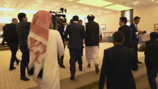 Des membres d'une délégation, dont un homme portant un turban et une tenue traditionnelle, marchent dans un hall accompagnés par des journalistes et des caméramans. La scène montre une rencontre diplomatique ou politique impliquant des représentants des Talibans.
