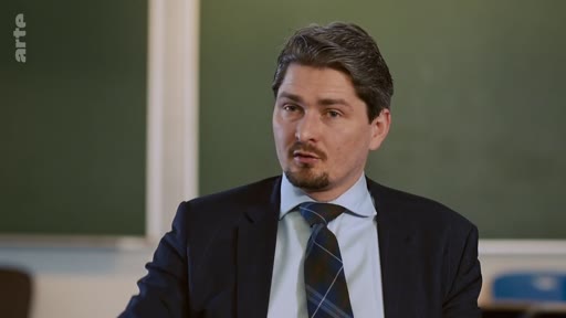 Un homme en costume sombre et chemise claire, portant une cravate, est filmé en plan moyen lors d'une interview devant un tableau vert. Il s'exprime face caméra dans un cadre formel.