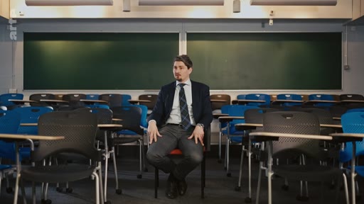 Un homme en costume-cravate est assis sur une chaise au centre d'une salle de classe vide, devant un tableau noir, lors d'une interview.
