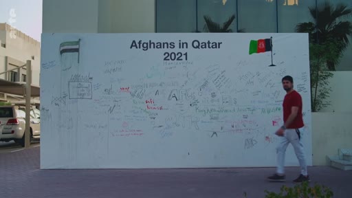 Un homme passe devant un grand panneau blanc extérieur portant l'inscription « Afghans in Qatar 2021 » et décoré de signatures et de drapeaux, illustrant la présence de réfugiés afghans au Qatar en 2021.