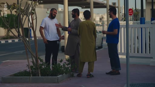 Quatre hommes, dont certains portent des vêtements traditionnels, discutent debout sur un trottoir dans une zone résidentielle en extérieur. La scène montre une interaction sociale informelle dans un environnement urbain moderne.