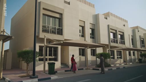 Des résidents, dont un homme et une enfant, se tiennent devant des maisons mitoyennes modernes dans un quartier résidentiel, probablement aux Émirats arabes unis. La scène montre l'architecture contemporaine des zones suburbaines avec des abris de voiture et des espaces extérieurs aménagés.