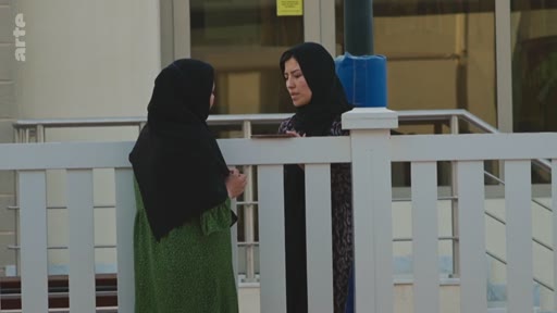 Deux femmes vêtues de hijabs, l'une en robe verte et l'autre en tenue sombre, ont une conversation devant une barrière blanche à l'extérieur d'un bâtiment. La scène illustre des interactions sociales dans un contexte culturel musulman.