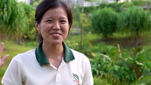 Une jeune femme en polo vert et blanc, probablement une employée des parcs nationaux, s'exprime devant la caméra dans un espace vert arboré. Elle participe à un reportage sur les initiatives de reforestation et la gestion des espaces naturels.