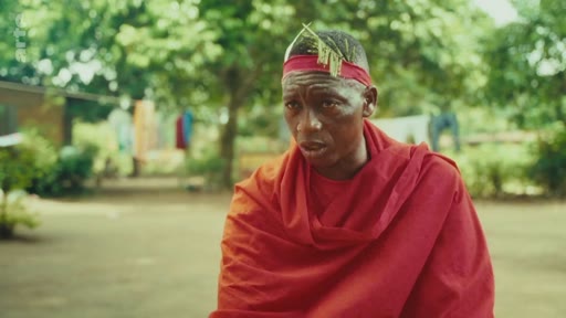 Un homme portant une tenue traditionnelle rouge et un ornement de tête s'exprime face caméra dans un cadre extérieur, illustrant les enjeux environnementaux et culturels locaux.