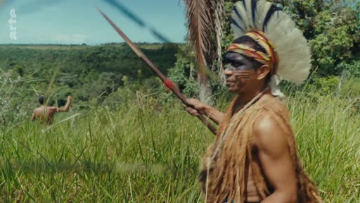 Un homme indigène portant une coiffe traditionnelle et des peintures corporelles tient une lance dans une zone de végétation dense, illustrant la lutte pour la préservation des terres et des cultures autochtones.