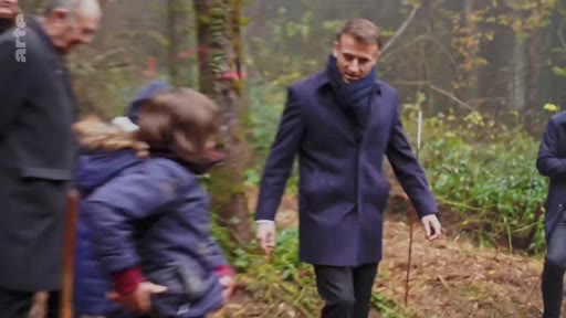 Le président français Emmanuel Macron marche dans une forêt lors d'une visite officielle consacrée à la reforestation, entouré d'enfants et d'autres personnes. Cette séquence illustre l'engagement présidentiel en faveur de la protection de l'environnement et de la sensibilisation des jeunes générations.