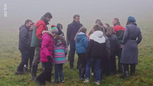 Le président français Emmanuel Macron échange avec un groupe d'enfants et des accompagnateurs dans un champ brumeux lors d'une initiative de plantation d'arbres. Cette séquence illustre l'engagement présidentiel en faveur de la reforestation et de la sensibilisation environnementale auprès des jeunes générations.